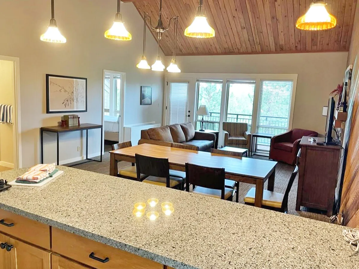 The image showcases a warm, inviting open-concept living space featuring a sleek granite countertop, a welcoming dining table perfectly set for gatherings, a cozy sofa area ideal for relaxing, and expansive windows that fill the room with natural light and offer views of the sunrise.