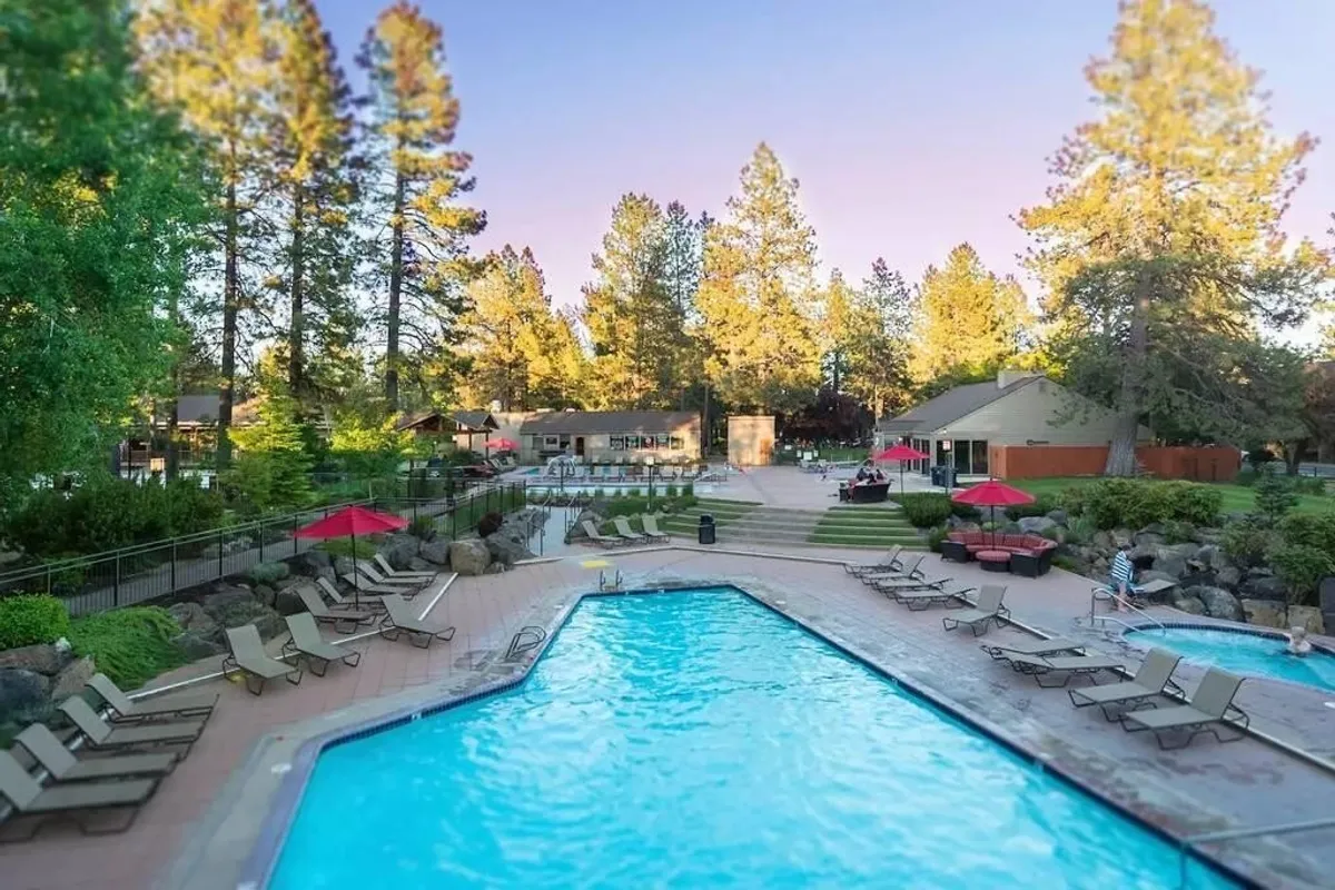 A tranquil outdoor swimming pool area with lounge chairs, cabana-style structures, and tall trees framing the space. The scene suggests both active swimming and relaxation, with ample space for socializing or sunbathing.
