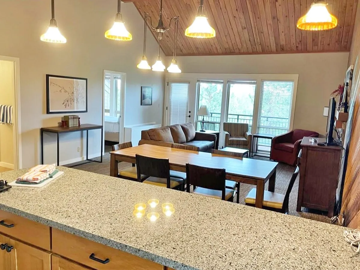 This image shows a welcoming open-plan living and dining area, featuring a granite countertop, a cozy dining table set for meals, a comfortable sofa, and large windows that let in abundant natural light. The space is ideal for family meals, social gatherings, and relaxing with a view of the outdoors.