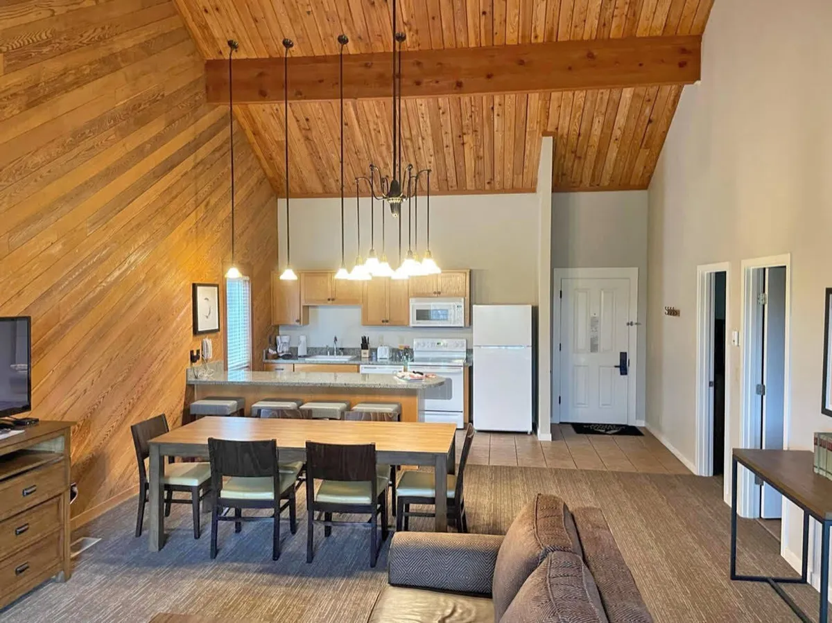 Featuring a spacious dining table and adjacent kitchen area, this image showcases wood-beam ceilings, a sofa, and an inviting, communal atmosphere. The layout suggests a seamless flow between cooking, eating, and relaxing—perfect for group meals and casual get-togethers.