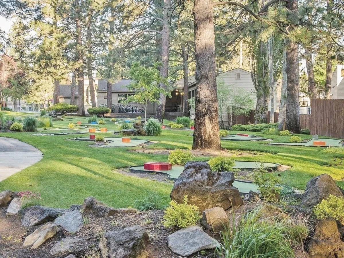 A cheerful mini-golf course set among lush greenery and stone pathways, with colorful obstacles and tall trees providing a playful, natural backdrop. The open, sunny space invites guests of all ages to enjoy outdoor recreation steps from their door.