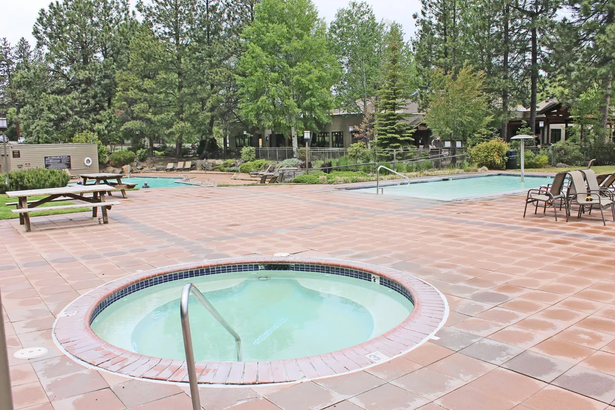 This image captures a sparkling swimming pool and inviting hot tub surrounded by landscaped greenery and picnic benches. The serene outdoor setting is ideal for relaxation, socializing, or enjoying a peaceful moment beneath the trees. The area is bathed in gentle natural light, emphasizing the tranquil resort atmosphere.