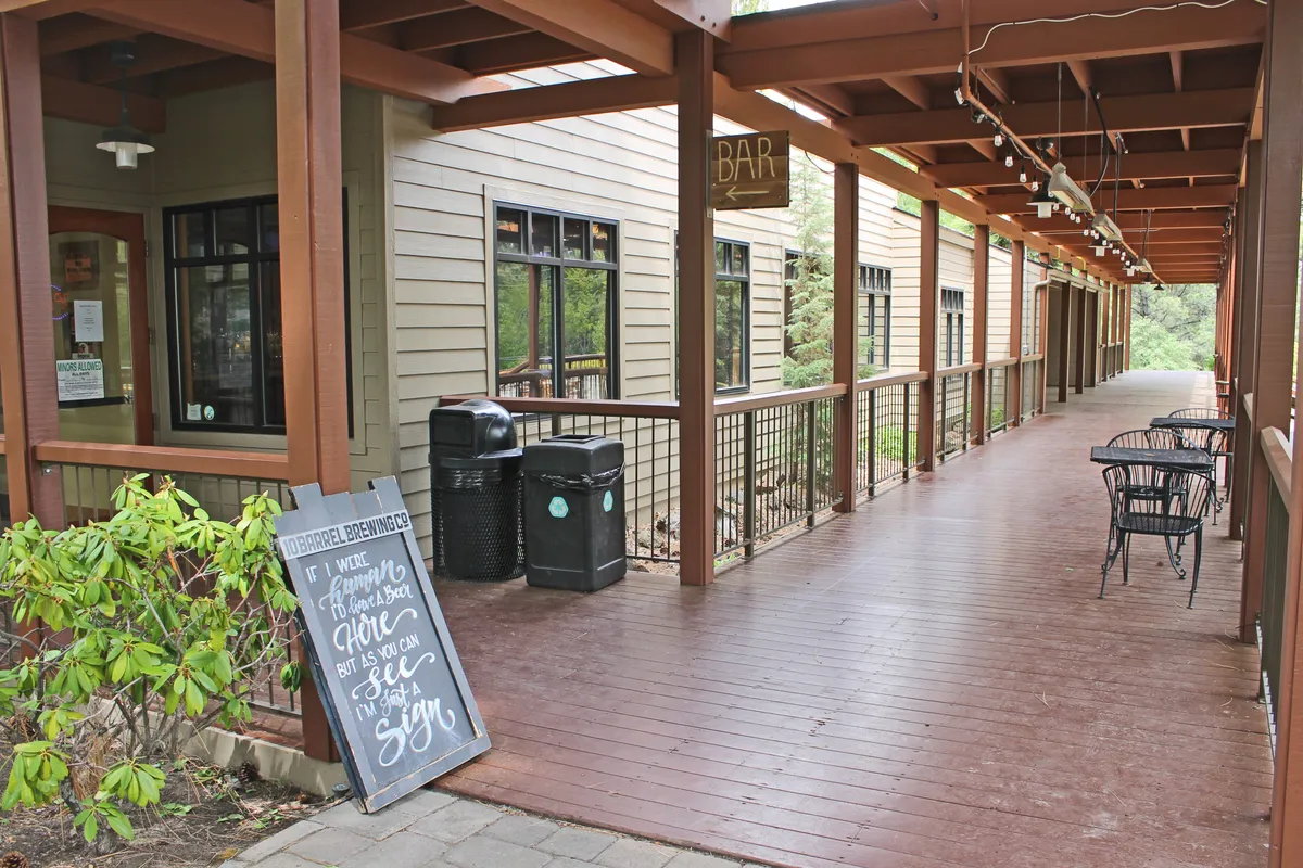 An inviting outdoor bar and seating area surrounded by lush greenery, with relaxed guests enjoying drinks under open skies. The scene features a well-maintained deck, prominent resort signage, and plenty of outdoor seating, evoking a sense of shared experiences and seasonal gatherings.