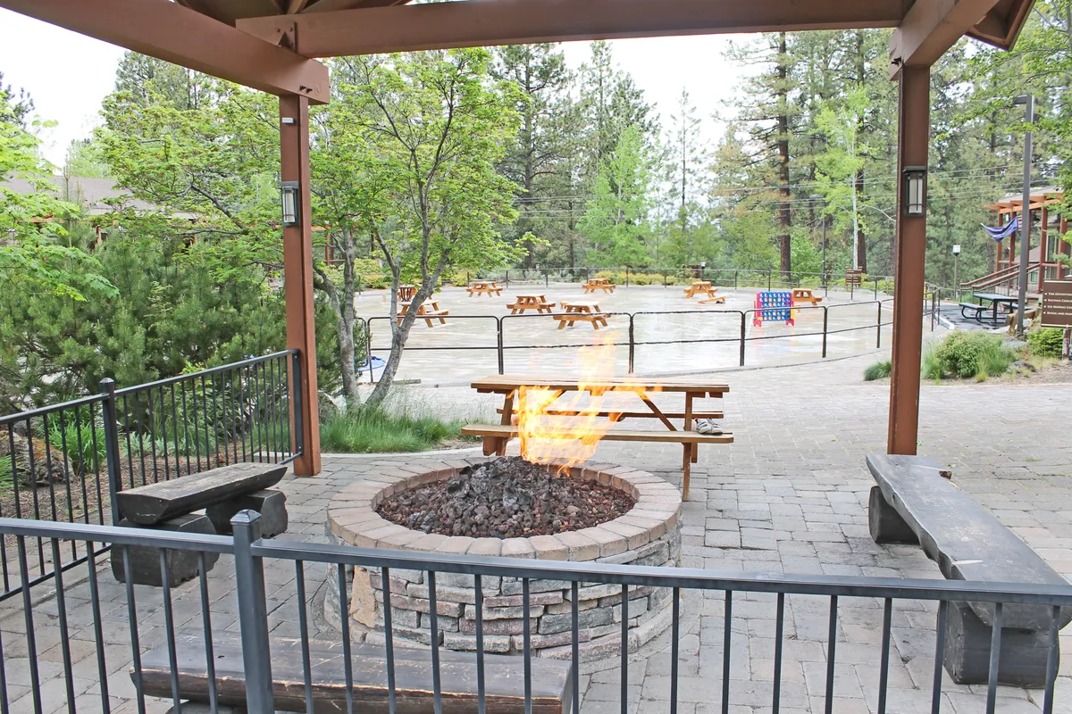 The photo showcases a communal outdoor fire pit area with surrounding benches and picnic tables in the background. The setup suggests a social, family- and pet-friendly atmosphere where guests might gather in the evenings to relax, tell stories, or enjoy a cozy night outdoors after a day of adventure.