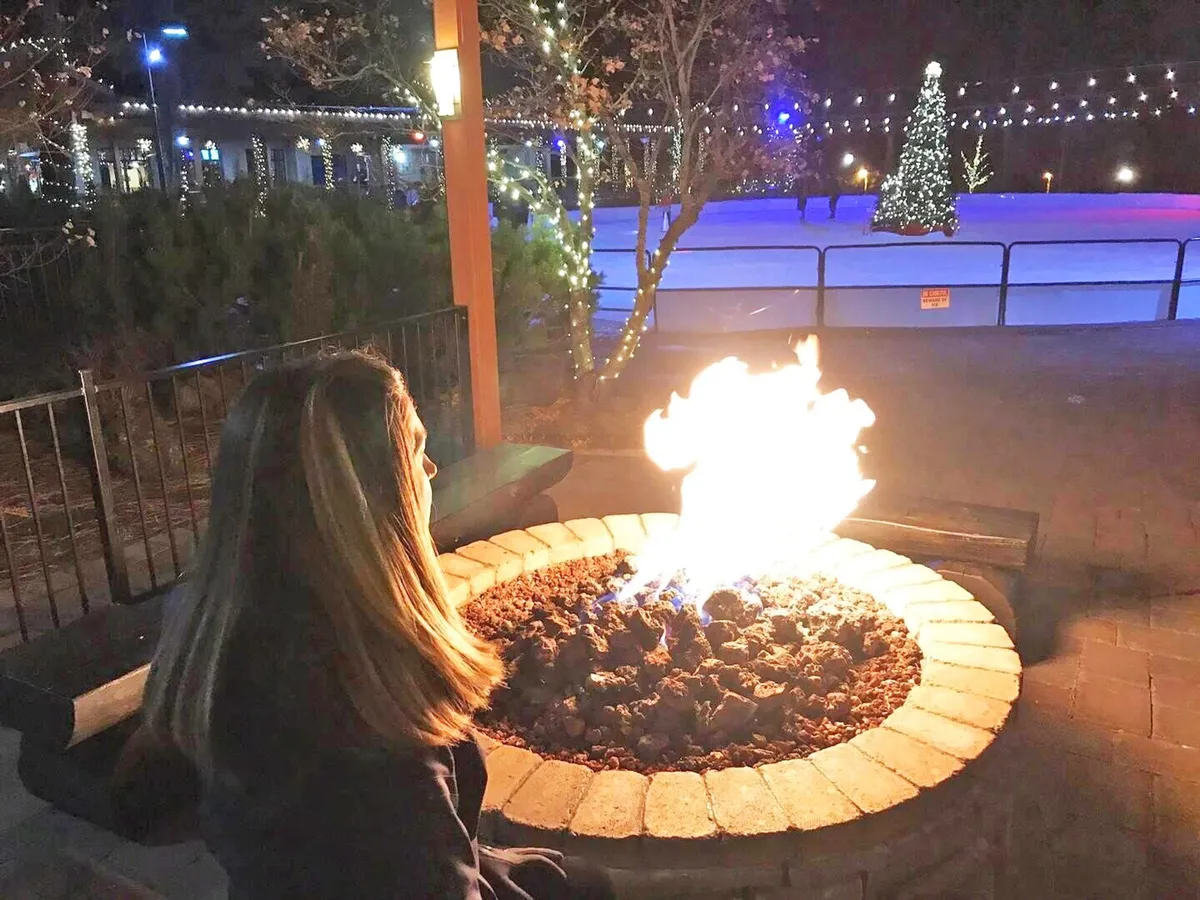 A lively winter scene featuring a glowing fire pit with someone seated nearby, surrounded by festive lights and a decorated Christmas tree. The outdoor ice skating rink is visible in the background with people skating, all set against a wintery backdrop.