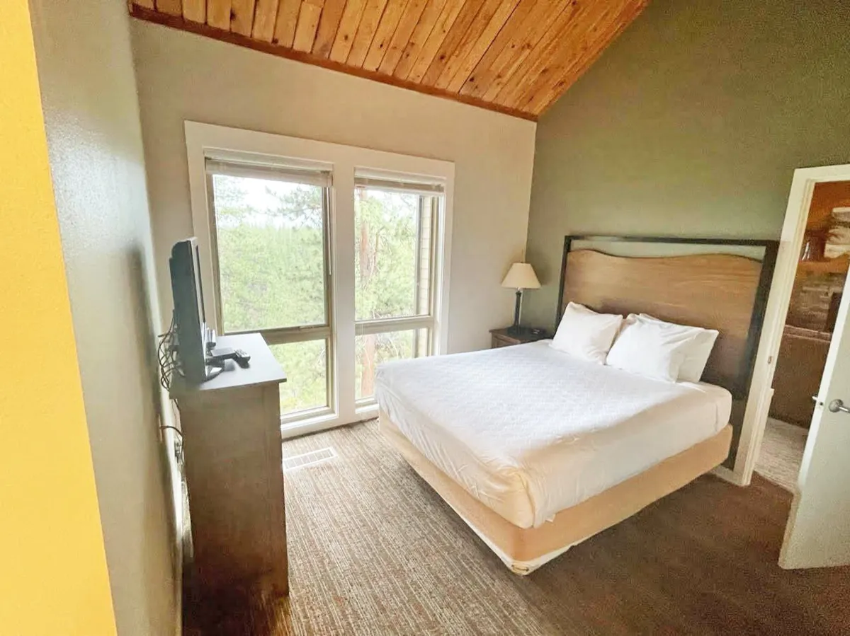 A cozy bedroom with a plush bed, large windows bringing in natural light, a TV, and a nightstand. The peaceful setting hints at a space perfect for relaxation, reading, or enjoying a quiet evening movie.